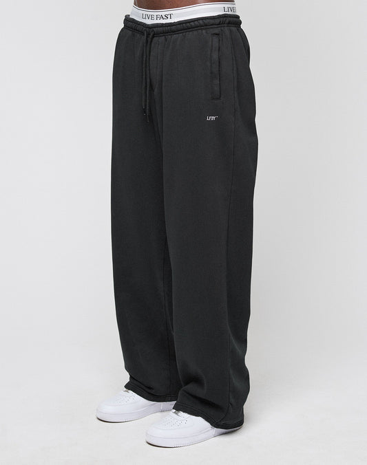 A person wears the LIVE FAST DIE YOUNG Basic Wide Jogger in black with a drawstring waist, white sneakers, and visible LIVE FAST branded underwear. The background is plain and light-colored.