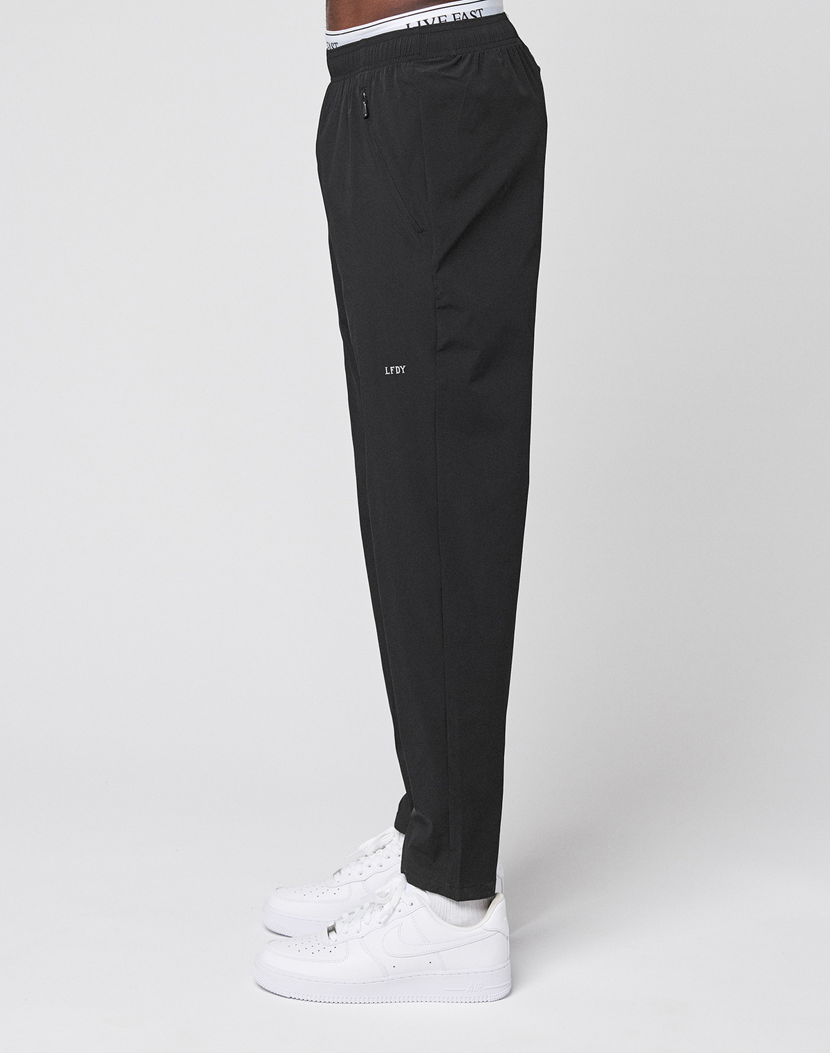 A person stands in profile against a plain light background wearing LIVE FAST DIE YOUNG LFDY Performance Pants in black with an oversize fit and LFYT branding, paired with white sneakers and a white waistband featuring LFYT TASTE text.