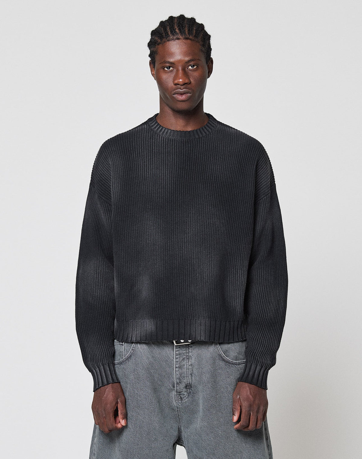 A person with braided hair wears the LIVE FAST DIE YOUNG LFDY Washed Knit Sweater, an oversized dark ribbed piece, paired with baggy gray 100% cotton jeans. They stand against a light gray background, facing forward with a neutral expression.