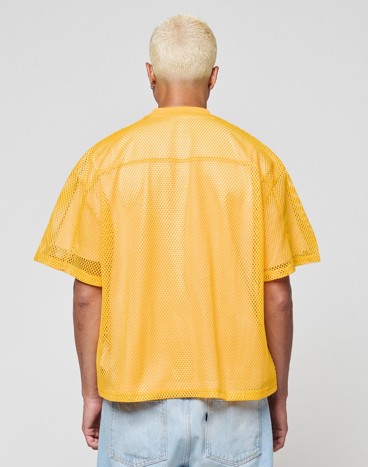 A person with short, bleached blonde hair is seen from behind wearing the LIVE FAST DIE YOUNG LFDY Boxy Jersey in yellow mesh and light blue jeans, set against a plain gray background.
