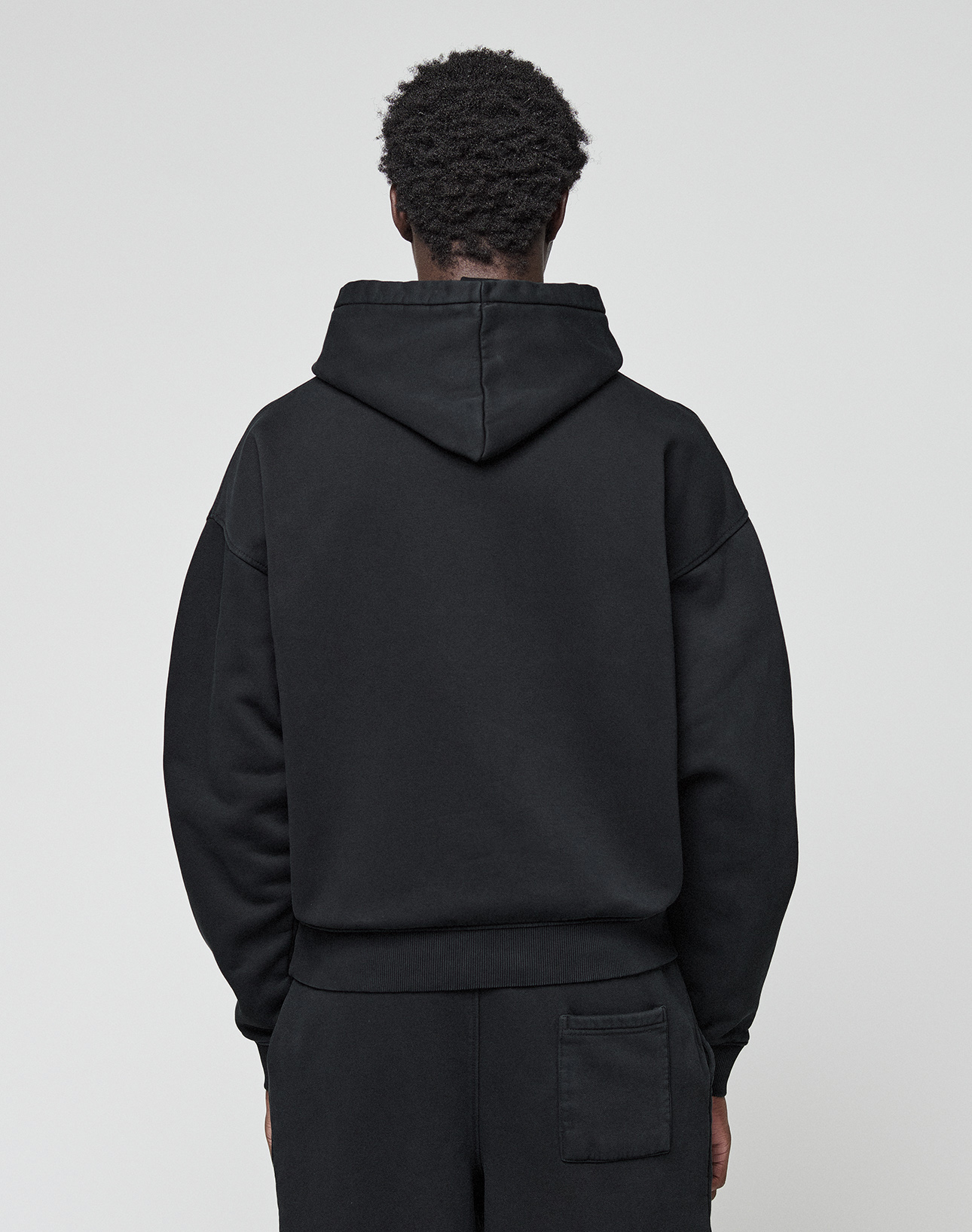 Seen from behind against a plain light background, a person with short curly hair wears the LIVE FAST DIE YOUNG Basic Livefast Hooded sweatshirt and matching black cotton pants in an oversize fit.