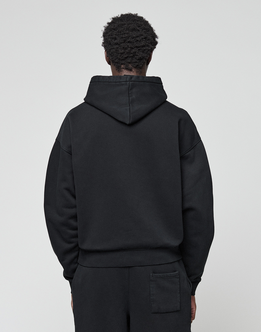 Seen from behind against a plain light background, a person with short curly hair wears the LIVE FAST DIE YOUNG Basic Livefast Hooded sweatshirt and matching black cotton pants in an oversize fit.