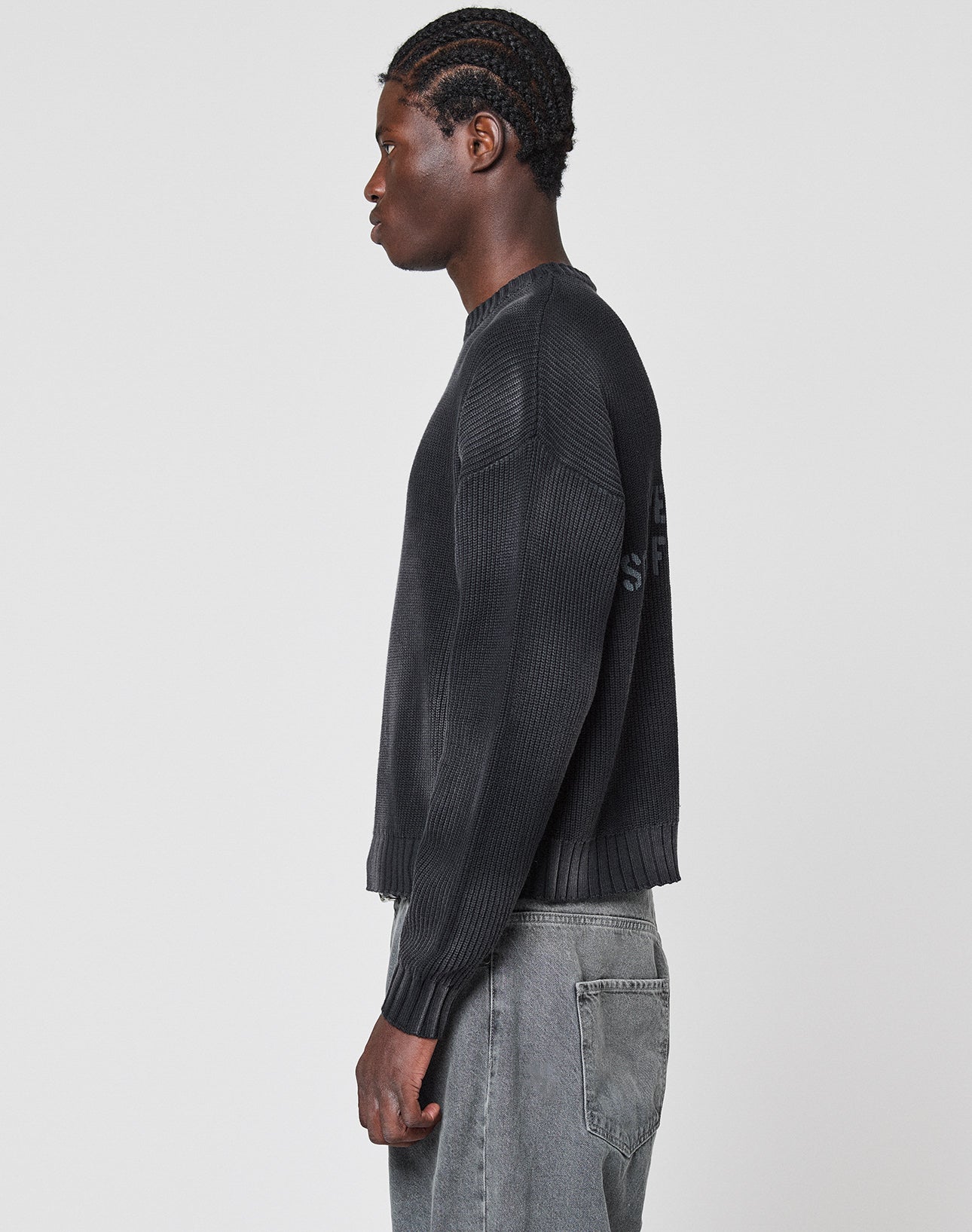 A man with braided hair stands in profile against a plain background, wearing the LFDY Washed Knit Sweater by LIVE FAST DIE YOUNG—an oversized, black ribbed sweater made from 100% Baumwolle—paired with gray jeans.