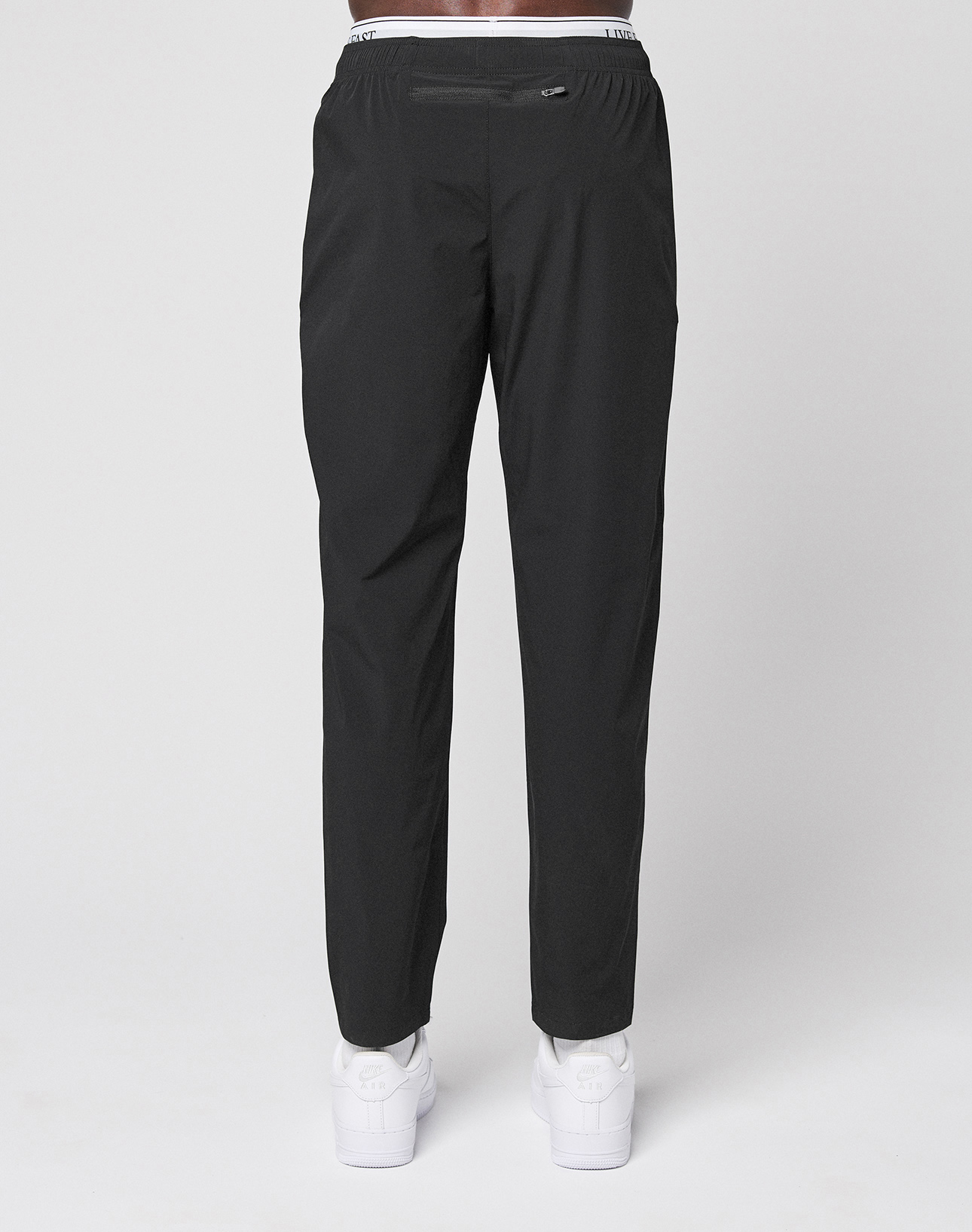 Someone stands facing away, wearing LFDY Performance Pants by LIVE FAST DIE YOUNG with an elastic waistband, branded details, and a reflective logo print, paired with white sneakers, against a plain light background.