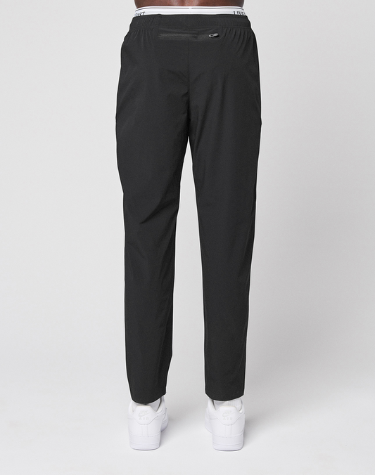 Someone stands facing away, wearing LFDY Performance Pants by LIVE FAST DIE YOUNG with an elastic waistband, branded details, and a reflective logo print, paired with white sneakers, against a plain light background.