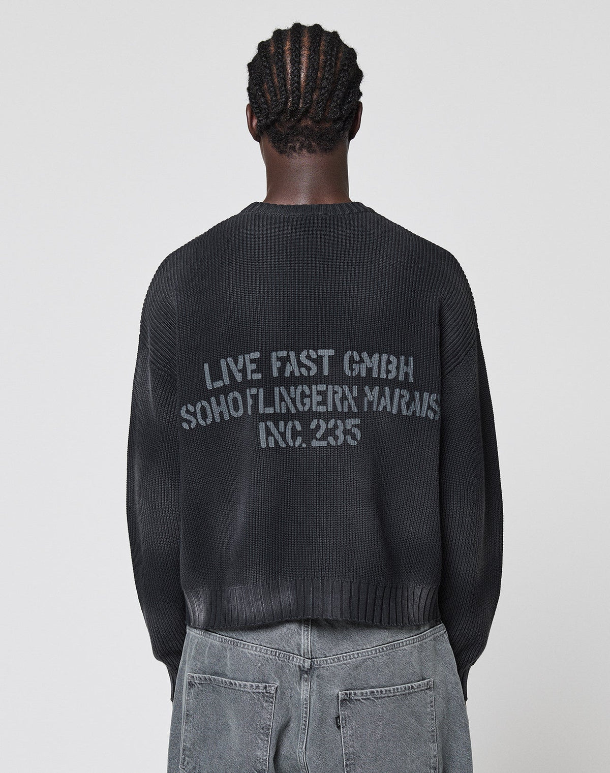 A person with braided hair, seen from behind in a plain light setting, wears gray jeans and the LIVE FAST DIE YOUNG LFDY Washed Knit Sweater—an oversize black 100% Baumwolle style with a bold gray backprint: LIVE FAST GMBH SOHO/FLINGERN MARAIS INC. 235. | model
