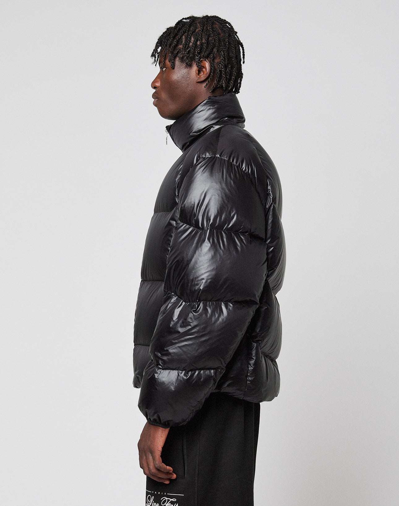 A person with short, braided hair stands sideways, wearing a shiny oversize LIVE FAST DIE YOUNG Live Fast Down Jacket with 600 fill power and black pants against a plain white background.