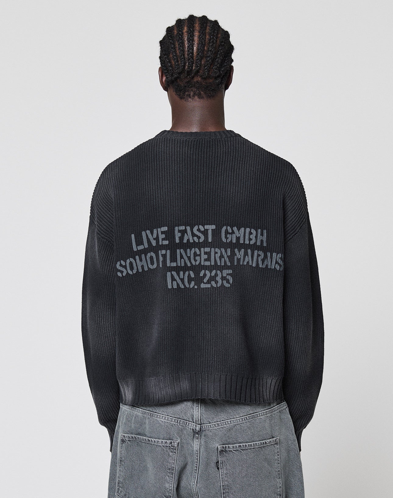 A person with braided hair, seen from behind in a plain light setting, wears gray jeans and the LIVE FAST DIE YOUNG LFDY Washed Knit Sweater—an oversize black 100% Baumwolle style with a bold gray backprint: LIVE FAST GMBH SOHO/FLINGERN MARAIS INC. 235.
