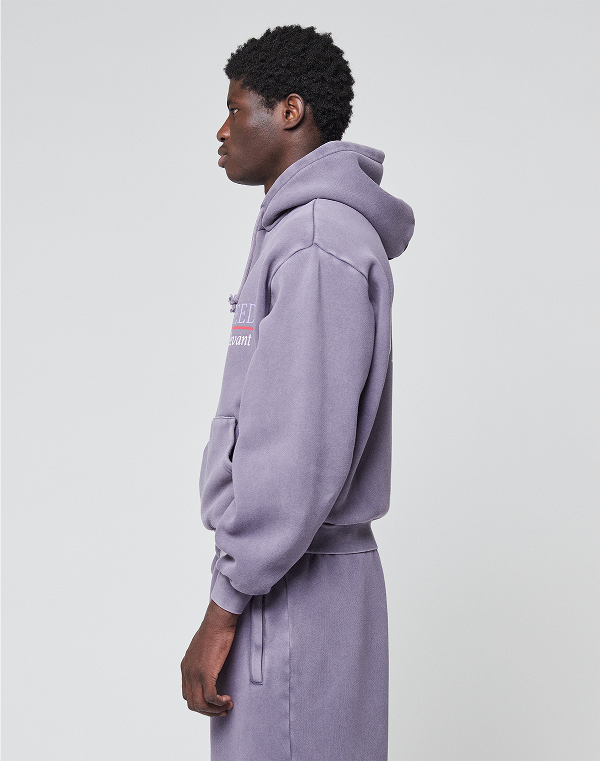 A person stands in profile wearing the LFDY Hotline Hooded Set by LIVE FAST DIE YOUNG, featuring a light purple hoodie with drawstring hood and oversize fit, hands by their sides against a plain light gray background.