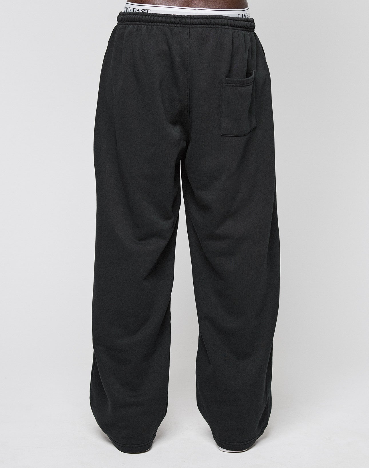 A person wears LIVE FAST DIE YOUNGs Basic Wide Jogger—loose-fitting black sweatpants with an elastic waistband, single back pocket, and oversized fit—while standing against a plain white background.