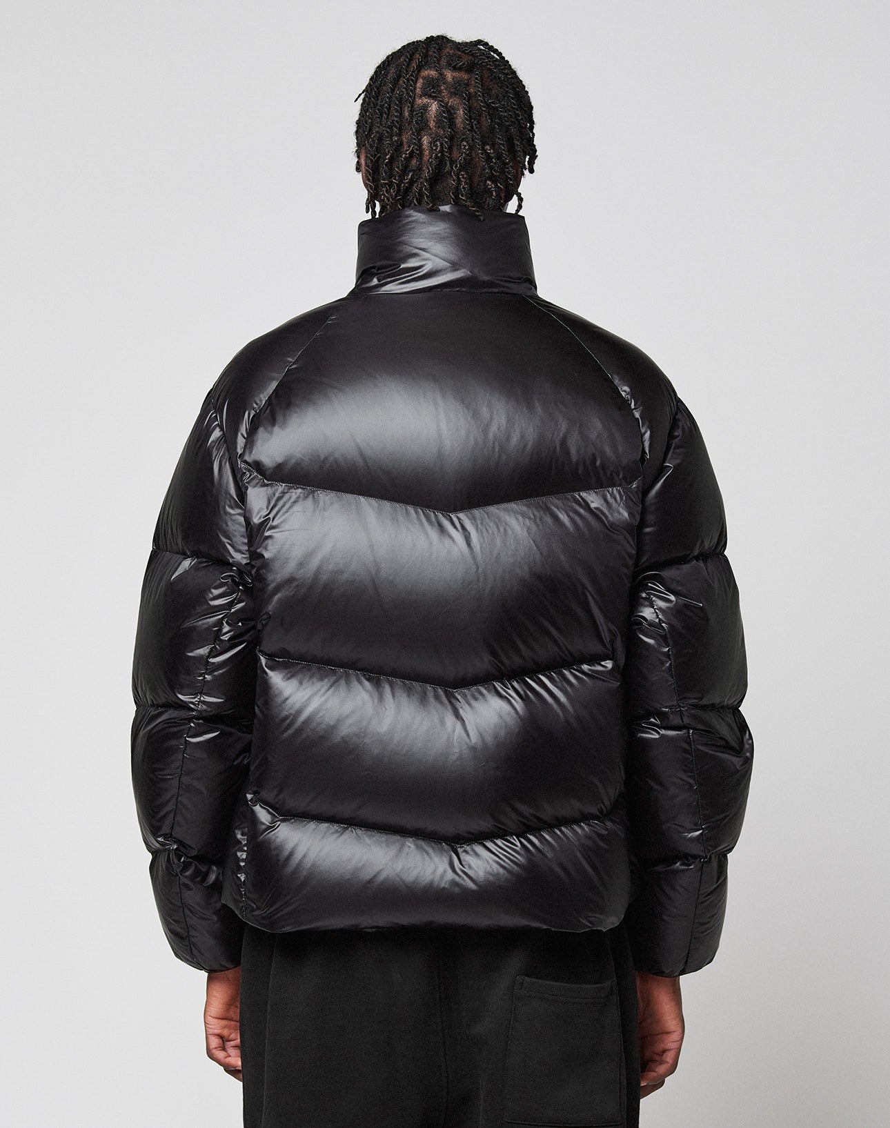 A person with short, twisted hair is seen from behind, wearing the Live Fast Down Jacket by LIVE FAST DIE YOUNG with 600er Fillpower and a shiny, wasserabweisend finish, against a plain light gray background.