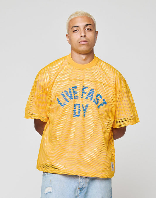 A person with short blond hair wears the LIVE FAST DIE YOUNG LFDY Boxy Jersey—a yellow, boxy mesh shirt with LIVE FAST D(Y) in blue—standing against a plain light gray background with a neutral expression.
