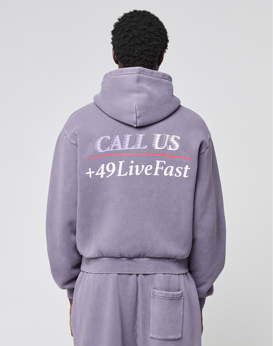 A person is shown from the back wearing an oversized lavender LFDY Hotline Hooded hoodie by LIVE FAST DIE YOUNG with drawstring hood, matching pants, and white CALL US +49LiveFast prints on front and back.