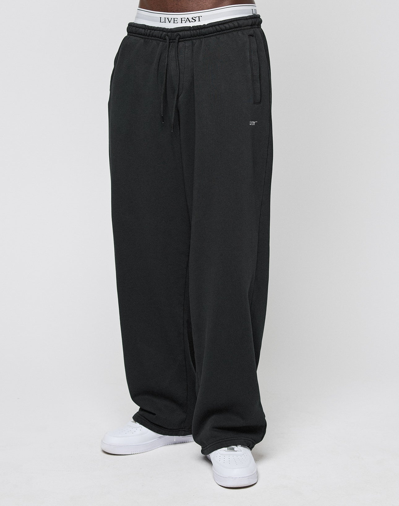 A person wears the LIVE FAST DIE YOUNG Basic Wide Jogger—loose-fitting black sweatpants with an elastic waistband featuring LIVE FAST text—paired with white sneakers, shown from the waist down against a plain white background.
