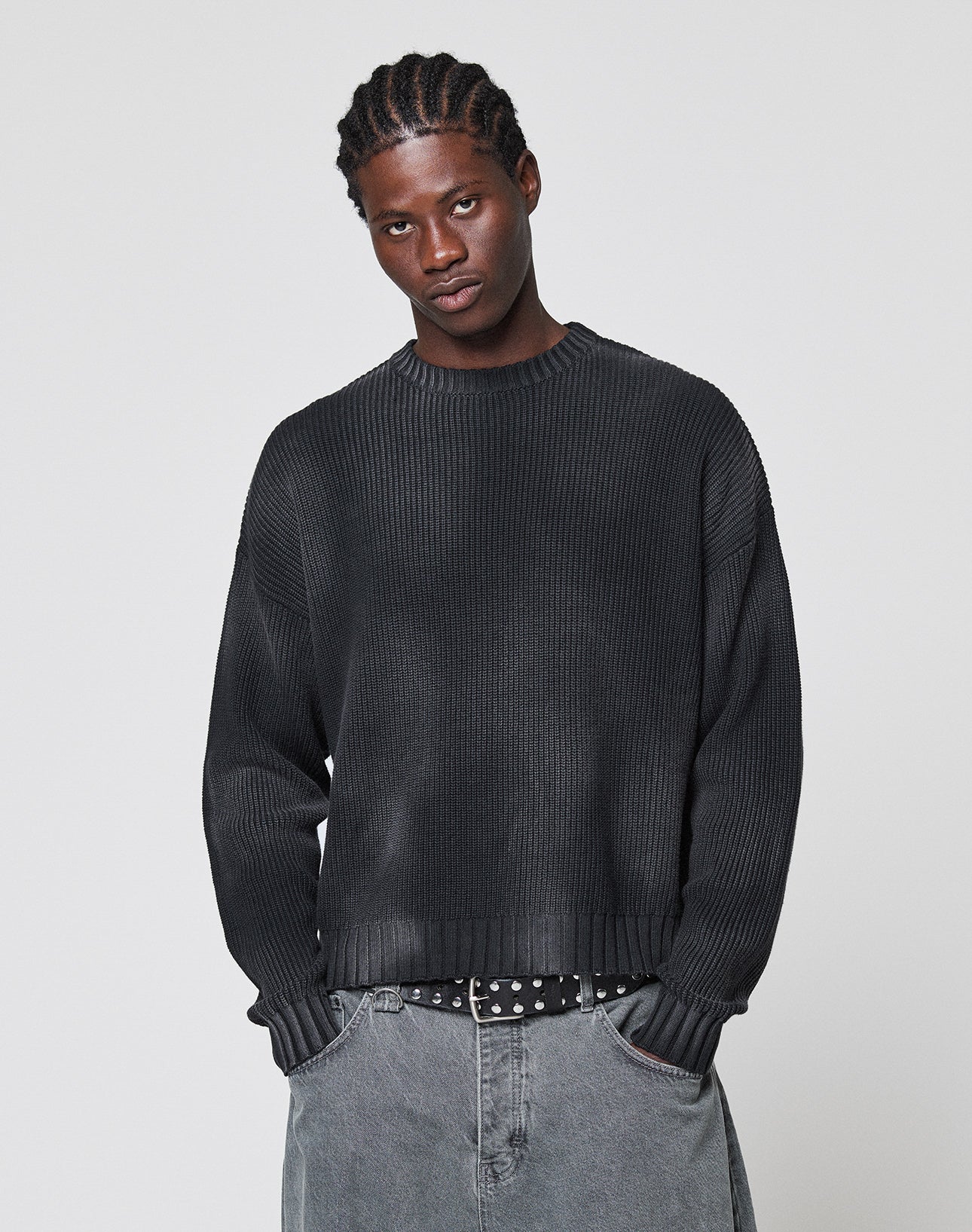 A young man with braided hair wears the LFDY Washed Knit Sweater by LIVE FAST DIE YOUNG and gray jeans. He stands against a plain light background, hands in pockets, looking slightly downward with a neutral expression.