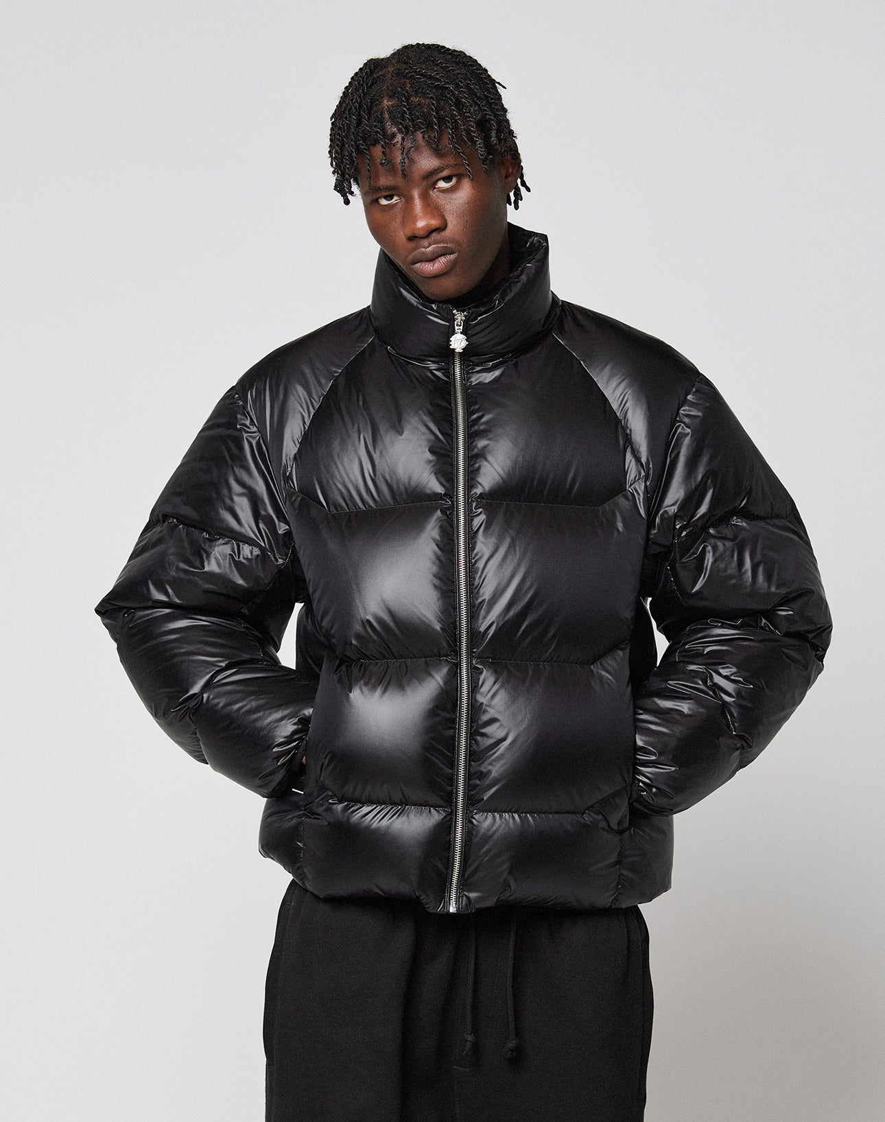 A person with short, twisted hair wears the LIVE FAST DIE YOUNG Live Fast Down Jacket—a shiny, wasserabweisende Oversize-Daunenjacke with 600er Fillpower—standing against a plain light gray background, hands in pockets, and a neutral expression.