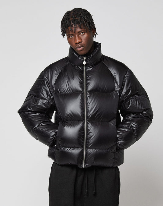 A person with short, twisted hair wears the LIVE FAST DIE YOUNG Live Fast Down Jacket—a shiny, wasserabweisende Oversize-Daunenjacke with 600er Fillpower—standing against a plain light gray background, hands in pockets, and a neutral expression.