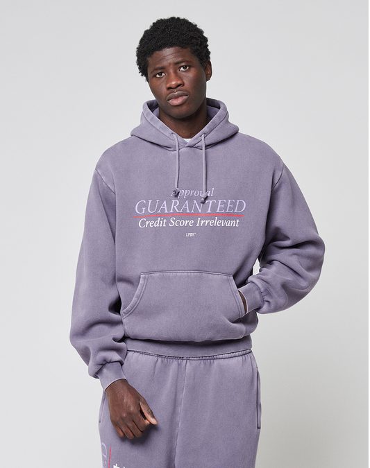 A young man wears a lavender LFDY Hotline Hooded by LIVE FAST DIE YOUNG with front and back print, featuring “Approval GUARANTEED Credit Score Irrelevant” and matching sweatpants, standing neutrally against a plain background.