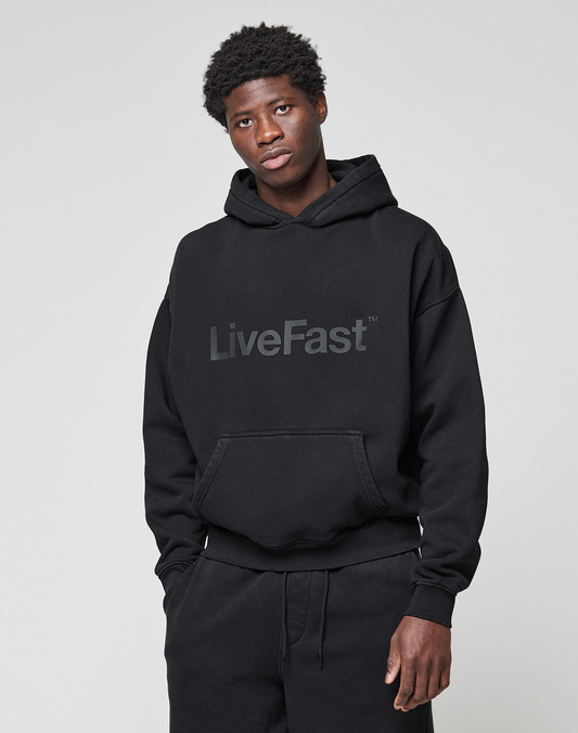 A young man models the Basic Livefast Hooded by LIVE FAST DIE YOUNG, featuring a gray "LiveFast" front print and oversize fit, standing against a plain light gray background with hands in his pockets, looking slightly to the side.