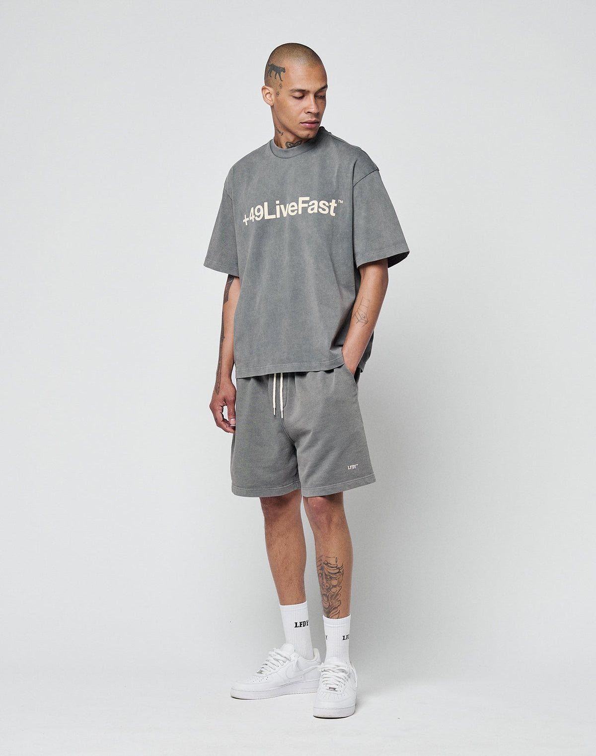 A man stands against a plain background wearing a Regular Fit gray #49LiveFast T-shirt, LIVE FAST DIE YOUNG Basic Sweat Shorts in gray with elastic waistband, white socks, and white sneakers. Tattoos are visible on his arms and leg.