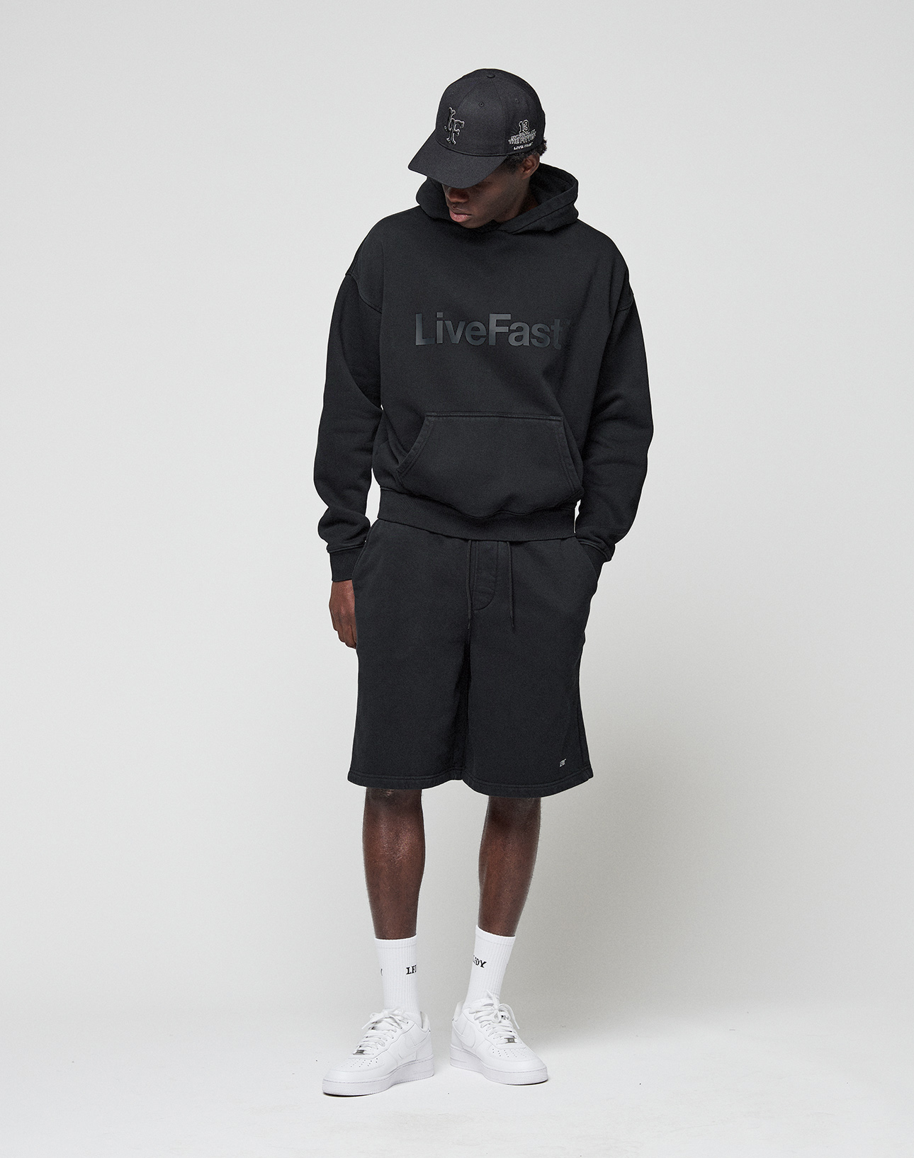 A person wears a black oversize fit Basic Livefast Hooded by LIVE FAST DIE YOUNG, paired with black shorts, black cap, white socks, and white sneakers. They stand against a plain light background, looking down with hands in pockets.