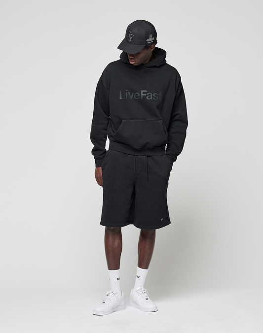 A person wears a black oversize fit Basic Livefast Hooded by LIVE FAST DIE YOUNG, paired with black shorts, black cap, white socks, and white sneakers. They stand against a plain light background, looking down with hands in pockets.