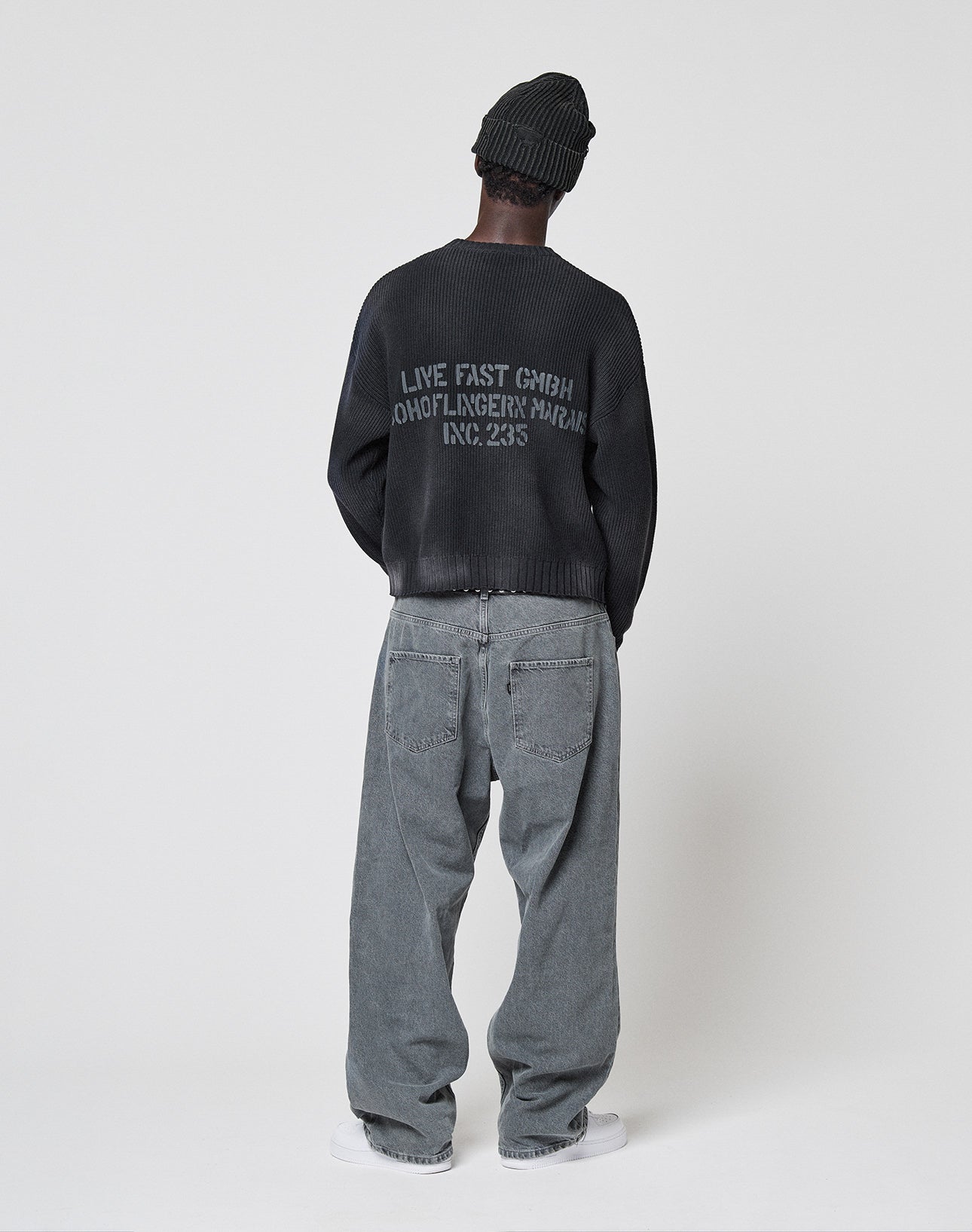 A person stands facing away, wearing a dark beanie, loose gray jeans, white sneakers, and the LIVE FAST DIE YOUNG LFDY Washed Knit Sweater with a backprint, set against a plain light background.