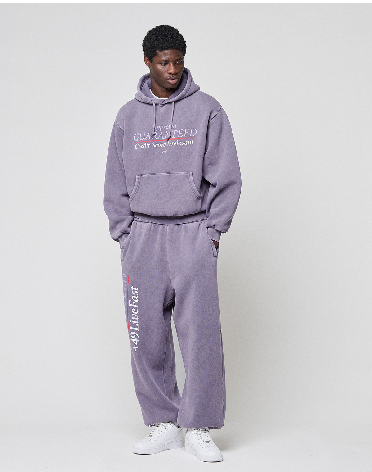A man wears the LIVE FAST DIE YOUNG LFDY Hotline Hooded—a purple, oversize hoodie with drawstring hood and bold front & back prints—paired with matching sweatpants and white sneakers, posing with a hand in his pocket against a plain background.