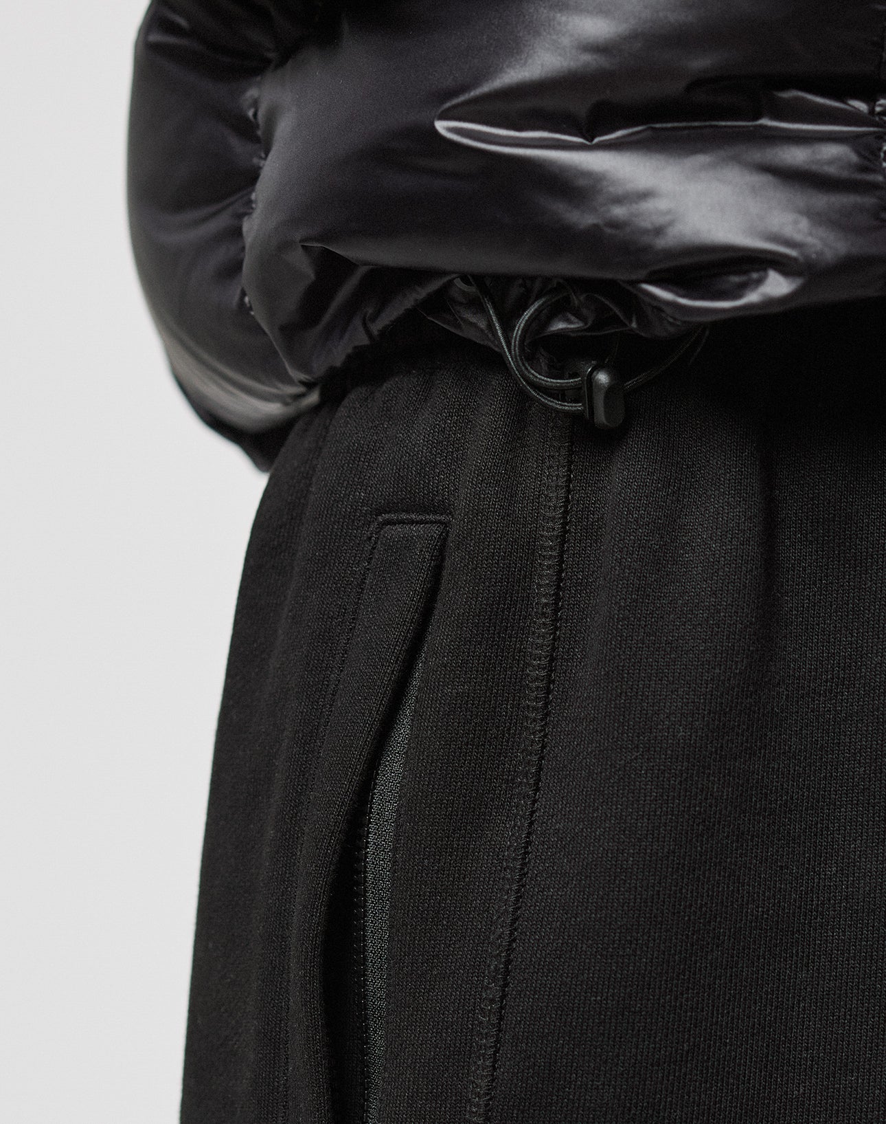 Close-up of a person wearing the LIVE FAST DIE YOUNG Live Fast Down Jacket in shiny black with 600er Fillpower and water-repellent fabric, paired with black pants featuring a visible zipper pocket and drawstring details.