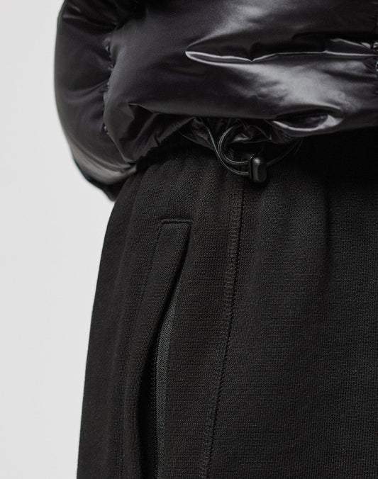 Close-up of a person wearing the LIVE FAST DIE YOUNG Live Fast Down Jacket in shiny black with 600er Fillpower and water-repellent fabric, paired with black pants featuring a visible zipper pocket and drawstring details.