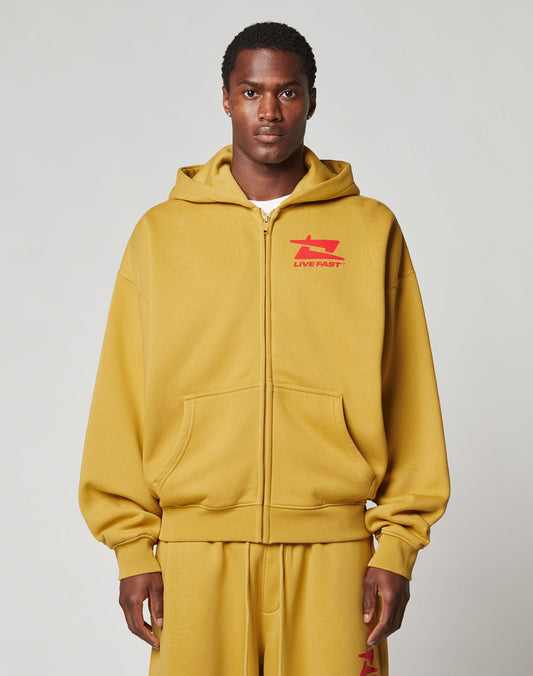 A man faces forward against a light gray background, wearing the LIVE FAST DIE YOUNG Live Fast Sweatsuit Jacket in mustard yellow with a bold LIVE FAST front print and red chest graphic, paired with matching oversized sweatpants.