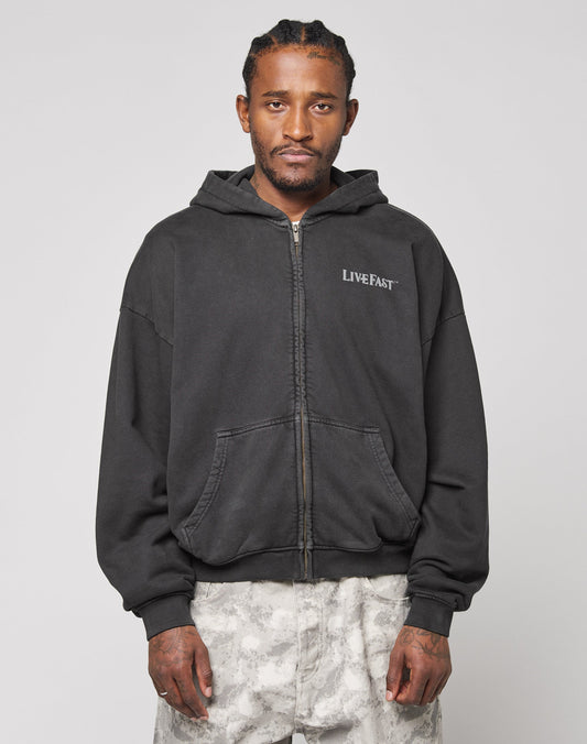 A man with braided hair wears the LIVE FAST DIE YOUNG LFDY Loyal Hooded Zip in black with LIVE FAST on the chest and light camo pants, posing against a plain gray background.