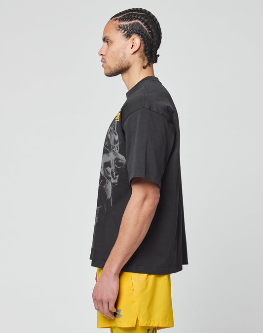 A man with braided hair stands in profile against a plain background, wearing the LIVE FAST DIE YOUNG LFDY Bark Tee—an oversize fit, vintage-wash black T-shirt made from 100% Baumwolle—paired with bright yellow shorts.