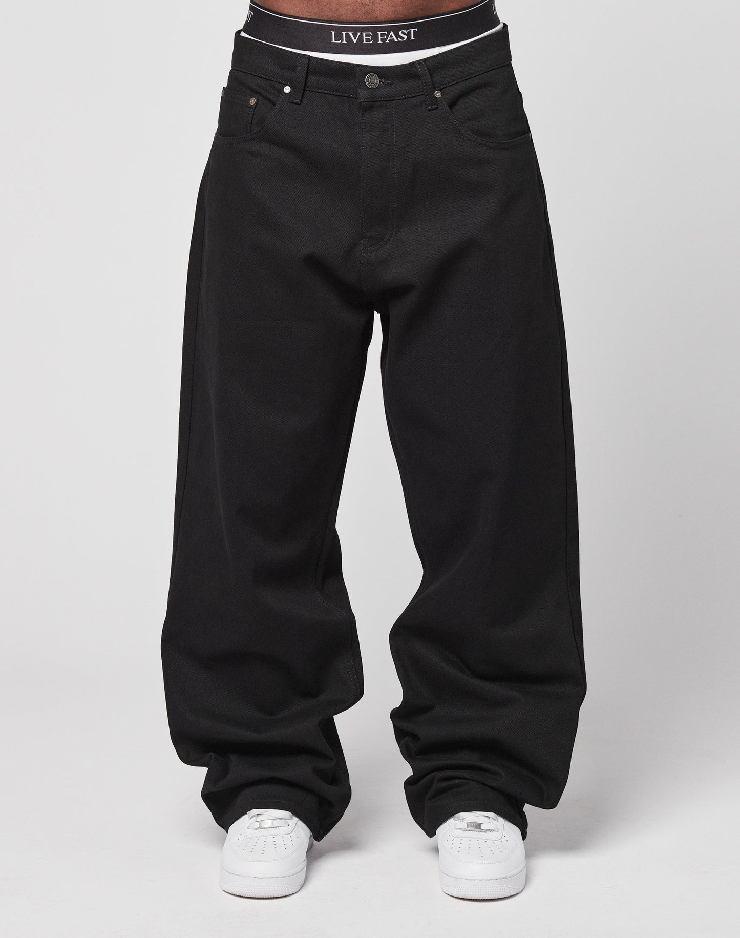 A person wears LFDY Baggy Denim in black with a baggy fit, paired with white sneakers. The waistband of their black underwear reads LIVE FAST. Made from 100% Baumwolle, the look is set against a plain white background.