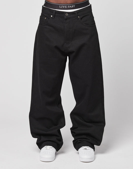 A person wears LFDY Baggy Denim in black with a baggy fit, paired with white sneakers. The waistband of their black underwear reads LIVE FAST. Made from 100% Baumwolle, the look is set against a plain white background.