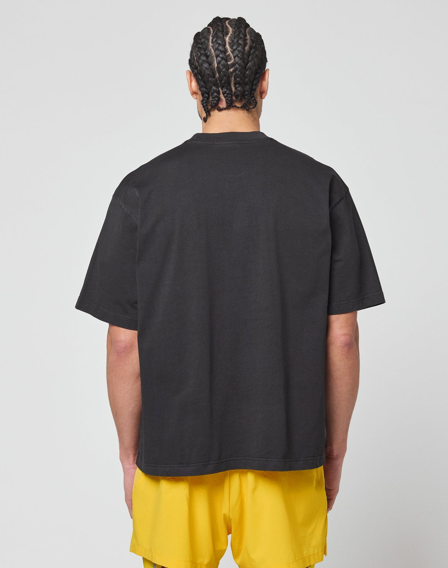 A person with braided hair faces away, wearing the LIVE FAST DIE YOUNG LFDY Bark Tee in black vintage wash and bright yellow oversized shorts, set against a plain white background.