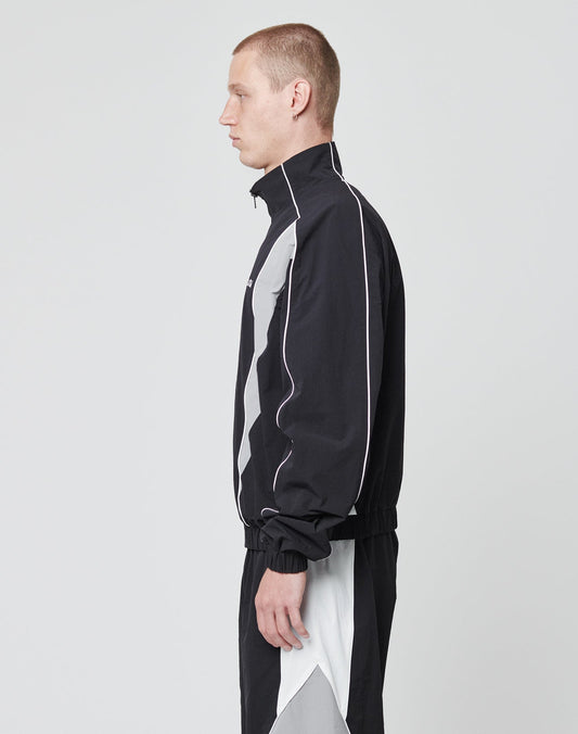 A person with short hair stands in profile, wearing the LIVE FAST DIE YOUNG Basic Nylon Trackjacket in black and gray with white piping, regular fit, against a plain light gray background.