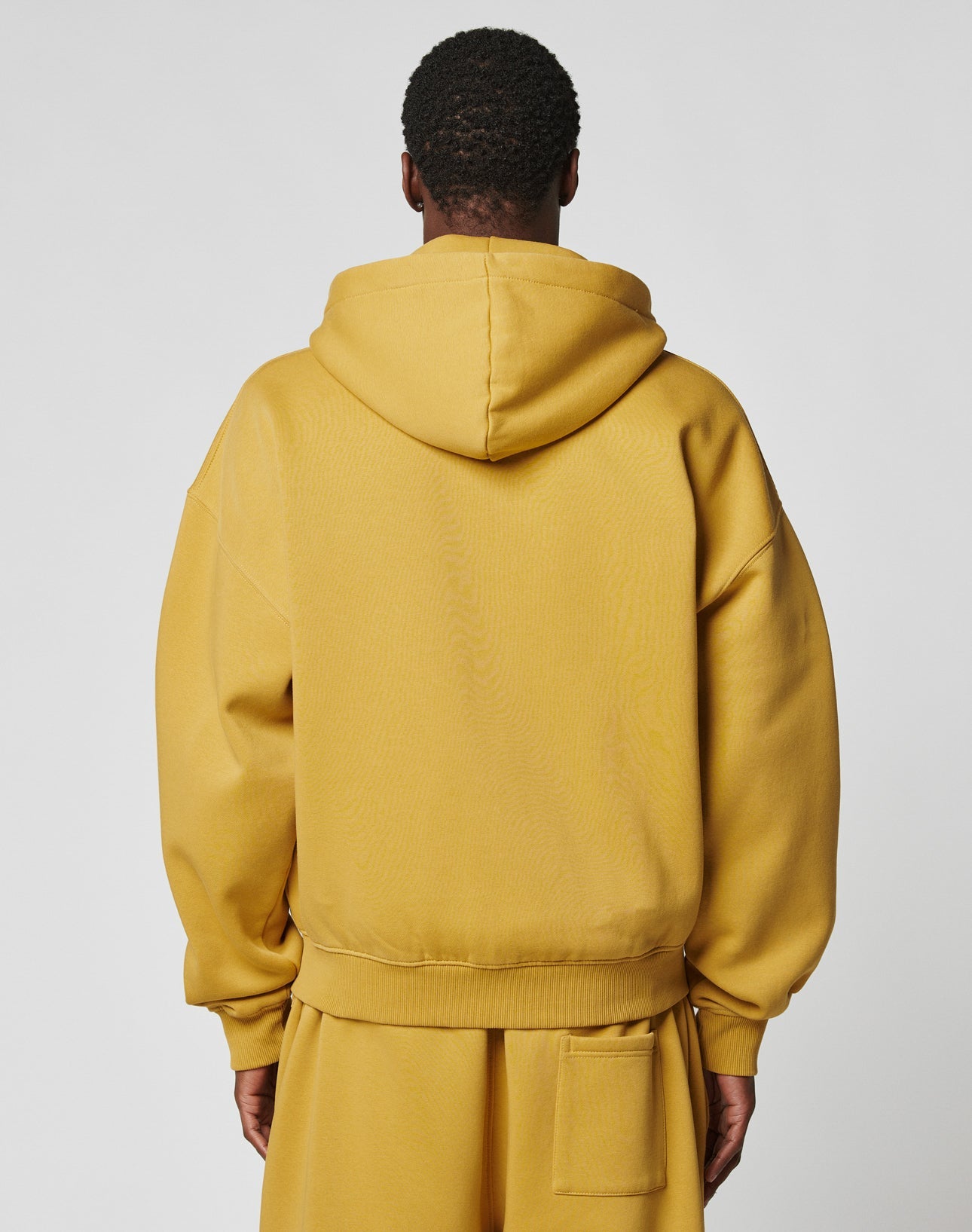 A person with short hair is seen from behind wearing the LIVE FAST DIE YOUNG Live Fast Sweatsuit Jacket in mustard yellow, featuring an oversized fit, large hood, and dropped shoulders. The background is plain and light-colored.