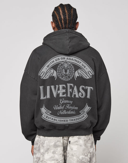 A person with braided hair, seen from behind, wears the LFDY Loyal Hooded Zip by LIVE FAST DIE YOUNG in an oversize fit. The black hoodie features large gray LIVE FAST text, vintage wash, decorative designs, and a circular emblem in 100% cotton.
