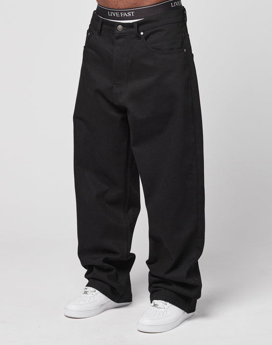 Someone wears LFDY Baggy Denim—baggy, loose-fitting black jeans made from 100% Baumwolle—with white sneakers and black LIVE FAST underwear visible at the waistband. The plain, light background highlights the outfit. | model