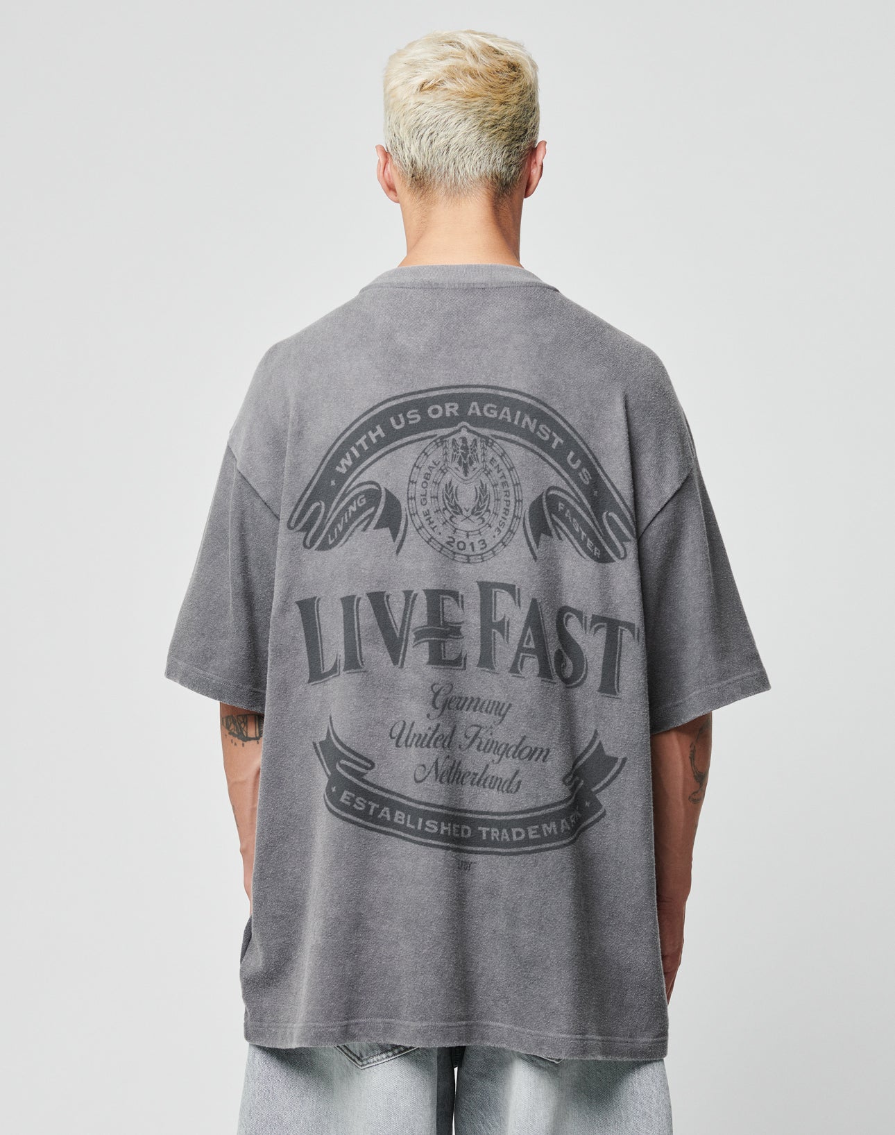 A person with short blond hair wears the LFDY Loyal Tee by LIVE FAST DIE YOUNG, a faded gray 100% Baumwolle oversize T-shirt with a large LIVE FAST graphic on the back, paired with light-wash denim shorts against a plain backdrop.