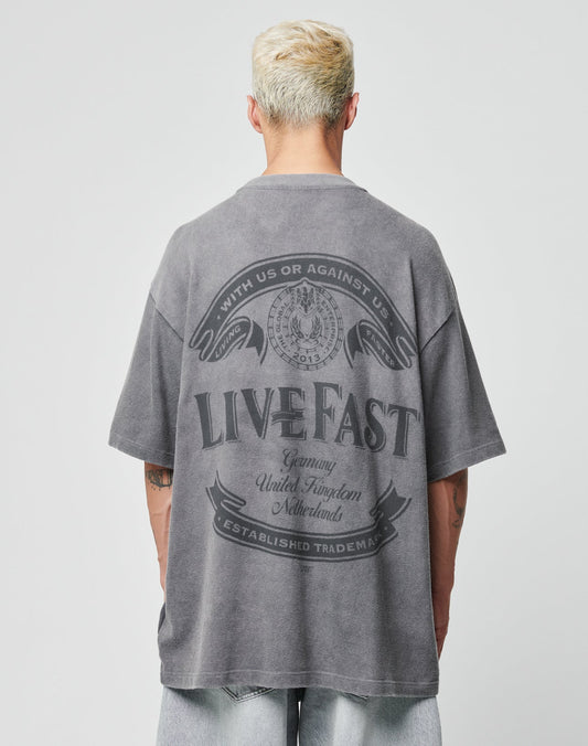 A person with short blond hair wears the LFDY Loyal Tee by LIVE FAST DIE YOUNG, a faded gray 100% Baumwolle oversize T-shirt with a large LIVE FAST graphic on the back, paired with light-wash denim shorts against a plain backdrop.
