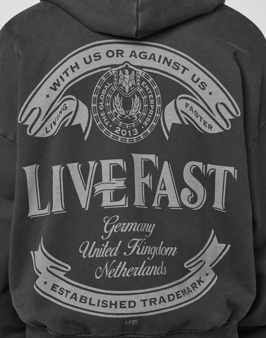 The LFDY Loyal Hooded Zip by LIVE FAST DIE YOUNG is an oversized, dark hoodie in 100% cotton with a vintage wash and bold back graphic—WITH US OR AGAINST US, LIVING FAST—with decorative banners listing Germany, UK, Netherlands.
