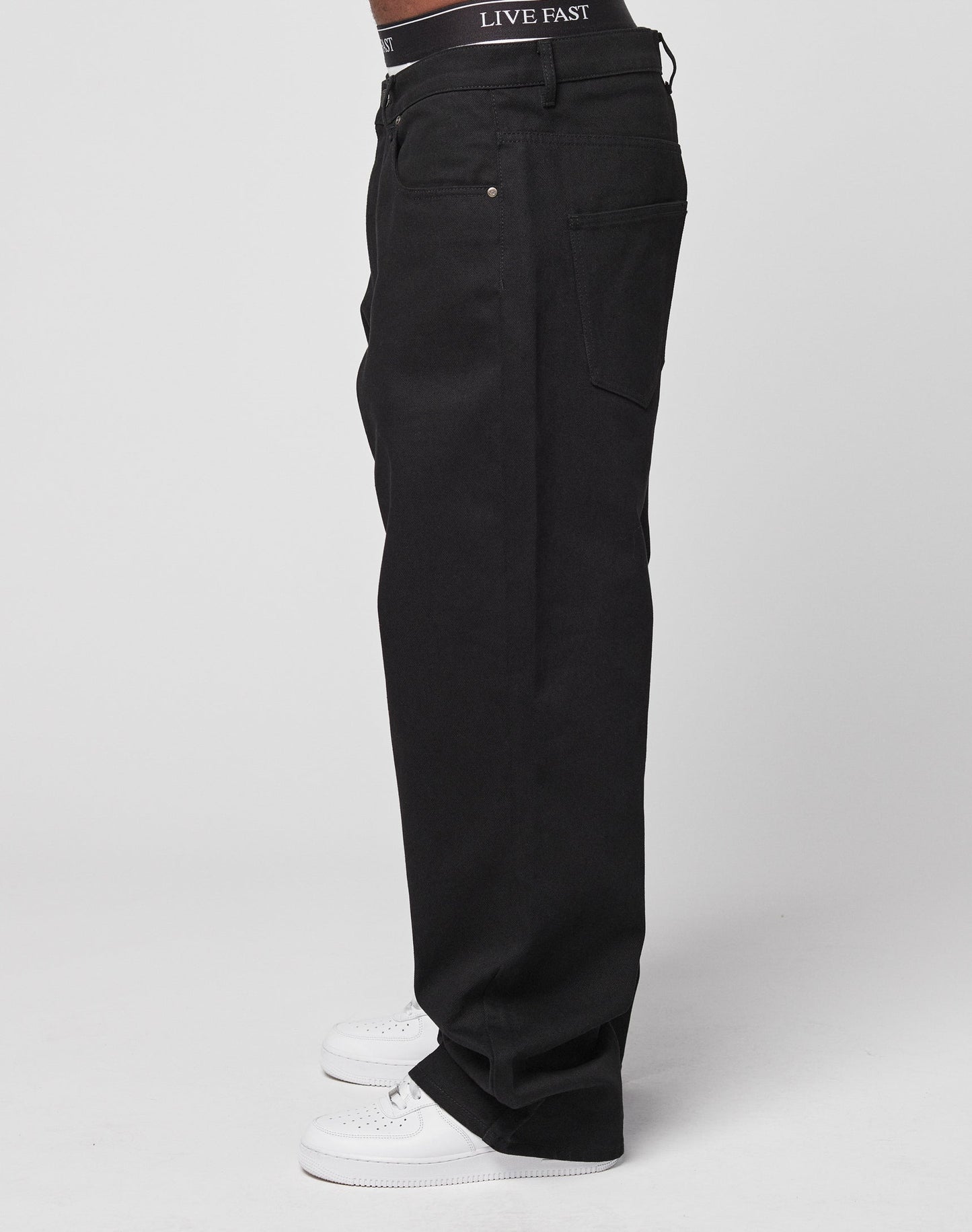A person wearing LFDY Baggy Denim black pants (100% Baumwolle) with a black LIVE FAST waistband and white sneakers, standing sideways against a plain white background.