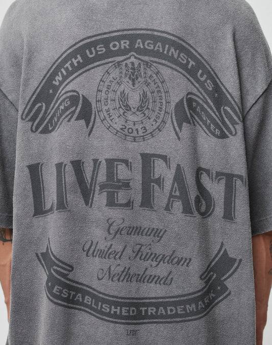 A person wears a gray LFDY Loyal Tee by LIVE FAST DIE YOUNG featuring “With us or against us. Living faster. The Good Life Established 2012. Live Fast.” Oversize fit, 100% Baumwolle, with Germany, UK, and Netherlands listed.
