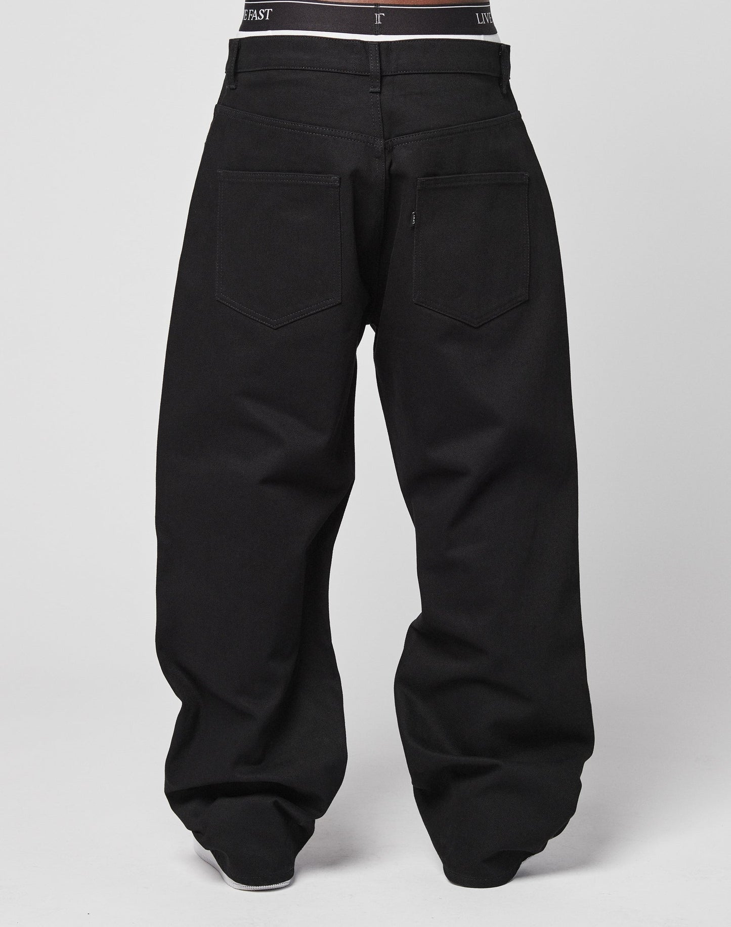 A person is seen from the back wearing LFDY Baggy Denim—loose-fitting black jeans with visible back pockets and a hint of black underwear at the waistband. Made from 100% Baumwolle. The background is plain and light-colored.