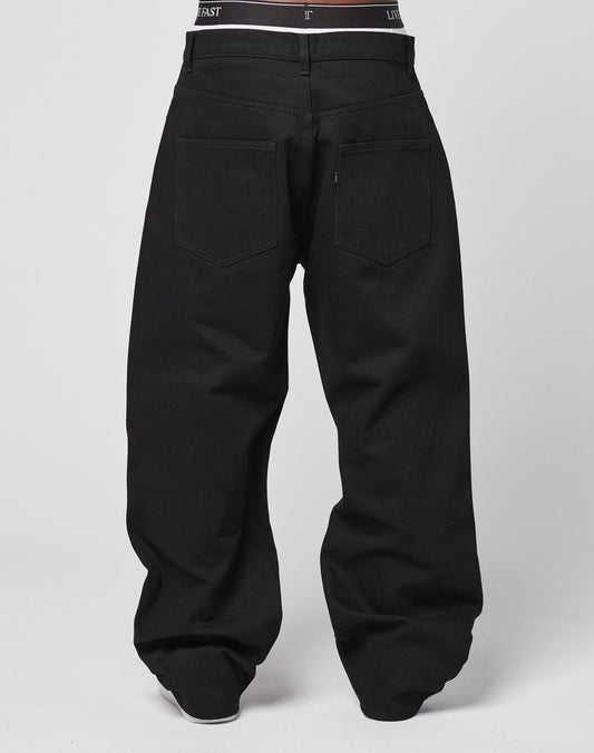 A person is seen from the back wearing LFDY Baggy Denim—loose-fitting black jeans with visible back pockets and a hint of black underwear at the waistband. Made from 100% Baumwolle. The background is plain and light-colored.