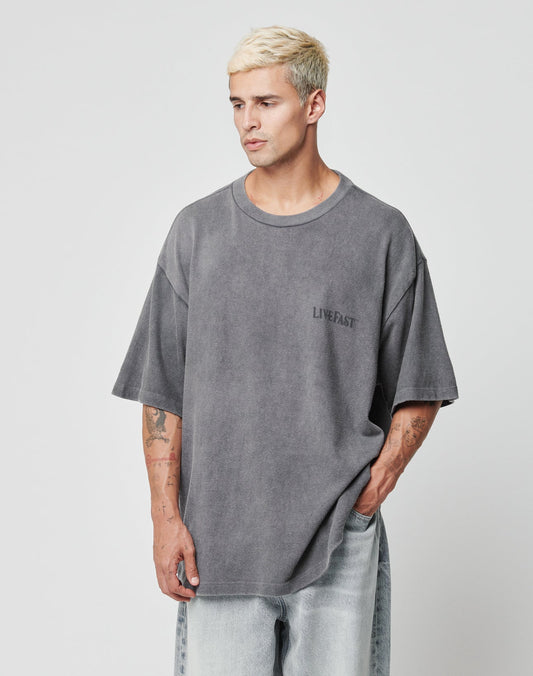 A man with short, bleached blonde hair wears an oversize faded gray LFDY Loyal Tee by LIVE FAST DIE YOUNG, made from 100% Baumwolle with LIVE FAST on the chest. He stands against a plain background, looking to the side with one hand in his pocket.