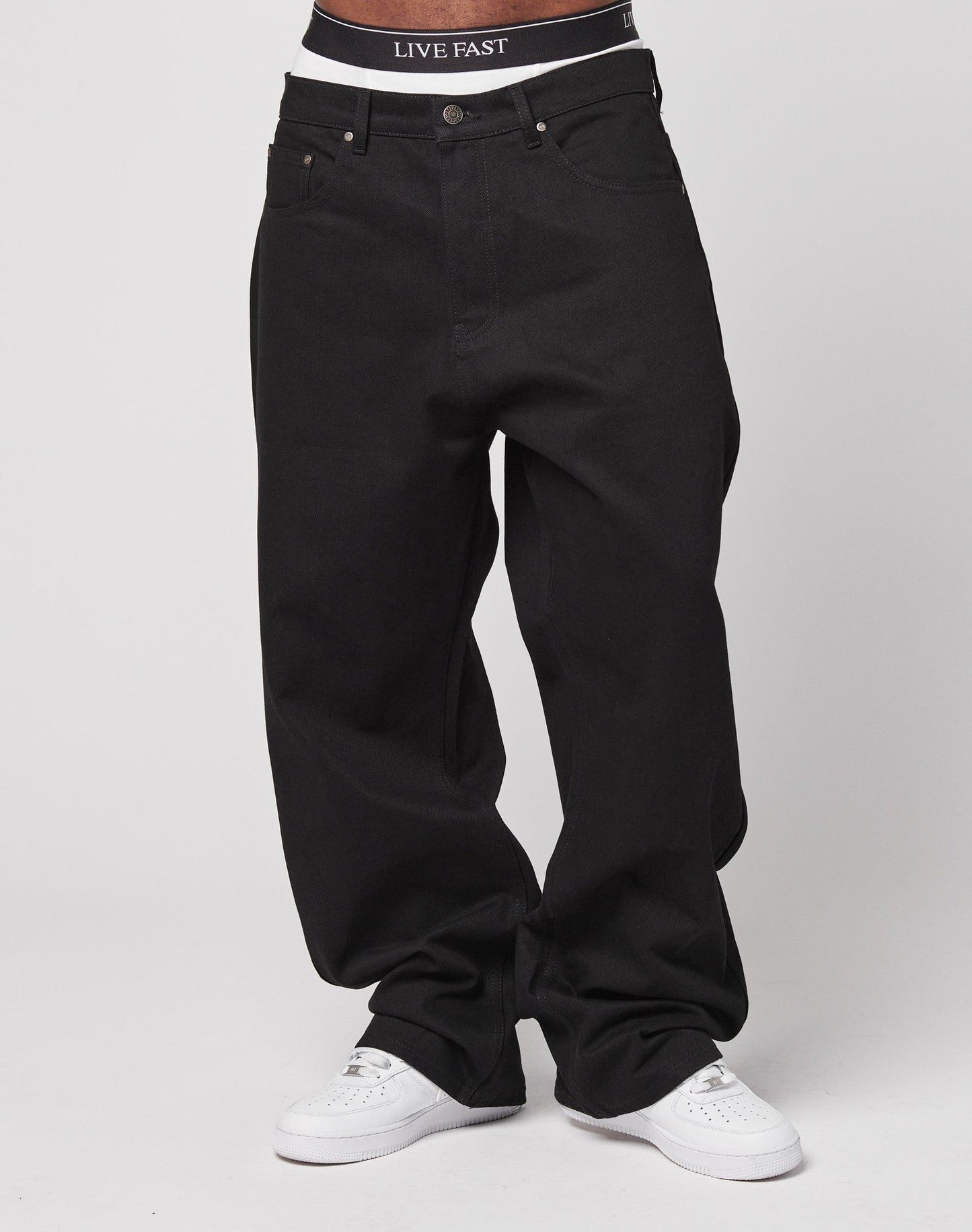 Model wears LFDY Baggy Denim by LIVE FAST DIE YOUNG in black 100% cotton, paired with white sneakers and visible black waistband featuring LIVE FAST text, set against a plain white background.