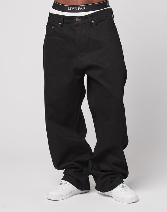 Model wears LFDY Baggy Denim by LIVE FAST DIE YOUNG in black 100% cotton, paired with white sneakers and visible black waistband featuring LIVE FAST text, set against a plain white background.